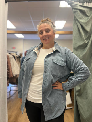 A woman wearing a light blue corduroy button-up shirt over a white t-shirt and black pants, standing in a fitting room.