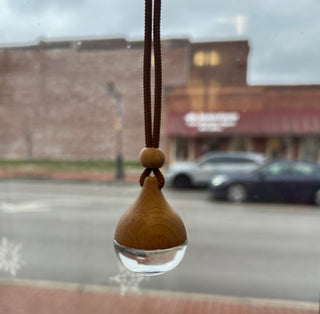 A wooden car air freshener with a clear glass bowl at the bottom and a brown cord, hanging in front of a window with a blurred street scene outside.