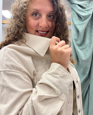 A close-up shot of a woman with curly light brown hair wearing a beige corduroy button-up shirt jacket, holding the collar up with one hand.