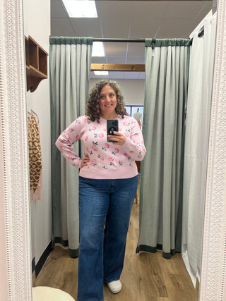 A woman takes a mirror selfie in a fitting room wearing a light pink long-sleeve crewneck sweater with a colorful floral pattern and blue jeans.