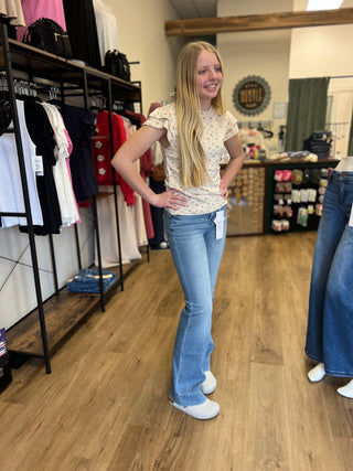 A woman with long blonde hair stands in a clothing store wearing a cream-colored short-sleeve top with a small floral pattern and light-wash blue flare jeans. She is smiling and posing with her hands on her hips, with clothing racks and wooden floors visible in the background.