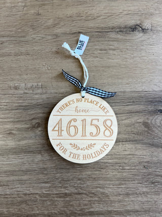 A round wooden Christmas ornament engraved with the number 46158 and the phrase 'There is no place like home for the holidays', featuring a black and white gingham ribbon bow and a small white tag on a wood-grain background.