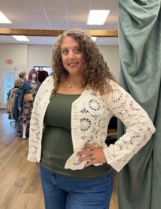 A woman with curly brown hair wearing a cream-colored, long-sleeve crochet cardigan over an olive green tank top and blue jeans, standing inside a boutique clothing store.