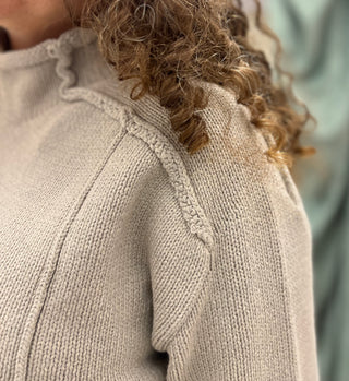 Close-up of a person wearing a chunky knit sweater in a warm light beige or oatmeal color, with curly brown hair visible at the neckline.