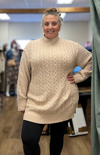 A woman wearing a cream cable-knit mock-neck sweater dress and black leggings, standing in a clothing boutique.