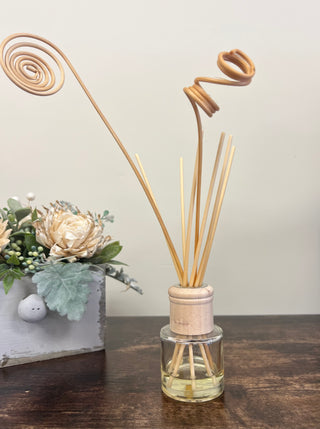 A reed diffuser with natural bamboo reeds and a light wood cap in a clear glass bottle, displayed on a dark wooden table next to a decorative floral arrangement.