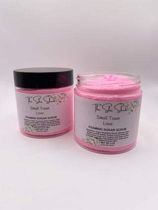 Two jars of The Shea Shield Small Town Love Foaming Sugar Scrub, with one jar closed showing a black lid and the other open showing the pink scrub inside, on a light gray background.
