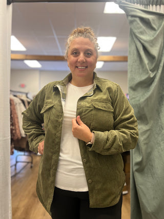 A woman wearing an olive green corduroy button-up shirt jacket over a white t-shirt and black pants, standing in a fitting room.