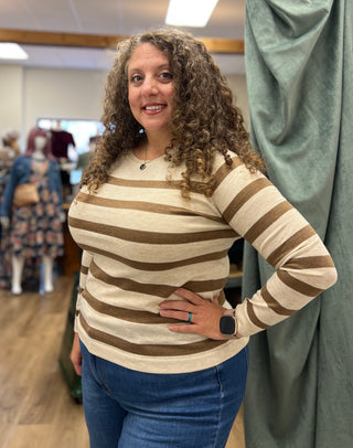 A woman with curly brown hair wearing a beige and brown horizontally striped long-sleeve knit top and blue jeans, standing in a boutique setting.