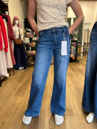 A person wearing medium-wash blue wide-leg jeans with a white price tag hanging from the waistband, standing in a clothing store with wood floors and racks of garments in the background.