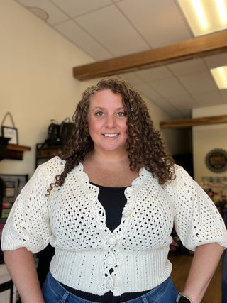 A woman with curly brown hair wearing a white crochet cardigan with puff sleeves over a black top and blue jeans, standing in a shop interior.