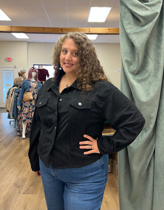 A woman with curly brown hair wearing a black denim jacket and blue jeans, standing in a clothing boutique with a green velvet curtain to her right.
