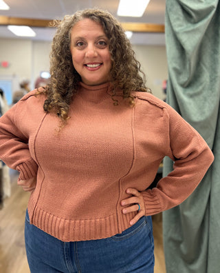 A woman with curly brown hair wearing a dusty rose ribbed knit mock neck sweater and blue jeans, standing in what appears to be a fitting room or studio.