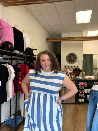 A woman wearing a blue and white striped dress, standing inside a clothing store with racks of garments and a mirror in the background.