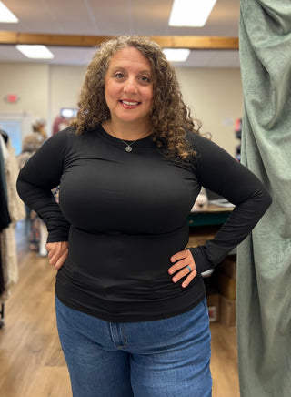 A woman with curly brown hair wearing a fitted black long-sleeve top and blue jeans, standing with hands on hips in what appears to be a clothing store or fitting room.