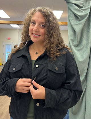 A woman with curly brown hair wearing a black corduroy button-up jacket over a green top, standing in an indoor fitting room or studio with a green curtain on the right.