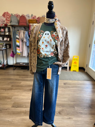 A mannequin dressed in a fall outfit featuring a green graphic t-shirt with a ghost and floral print, a brown crochet cardigan, wide-leg blue jeans, and a floral scarf, set in a boutique with wood floors.