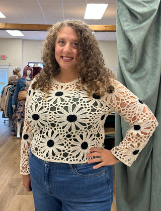 A woman with curly brown hair wearing a cream crochet long-sleeve top with black flower centers and blue jeans, standing in a clothing boutique.