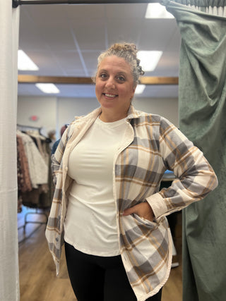 A woman modeling a long-sleeve plaid button-up shirt with a white t-shirt underneath, standing in a fitting room.
