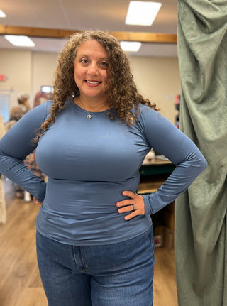 A woman with curly brown hair wearing a fitted, long-sleeve dusty blue top and blue jeans, standing in a fitting room or boutique with a green curtain on the right.