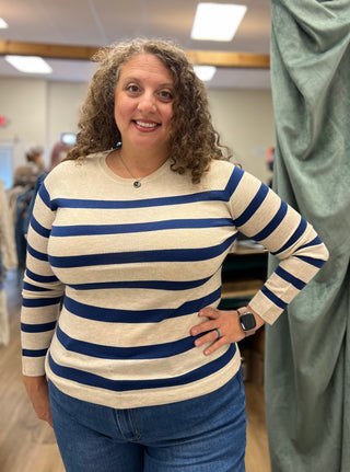 A woman with curly brown hair wearing a cream and navy blue horizontally striped long-sleeve knit top and blue jeans, standing in a clothing store.