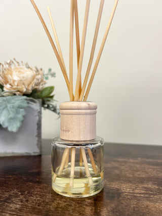 A clear glass reed diffuser bottle with pale yellow liquid and natural wood reeds, set on a dark wooden tabletop with a soft beige wall background.