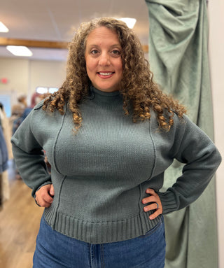 A woman with curly brown hair wearing a ribbed knit turtleneck sweater in a muted sage green, standing with hands on hips in what appears to be a boutique or fitting room.