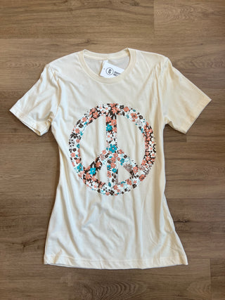 A cream-colored short-sleeve t-shirt laid flat on a wooden floor, featuring a large peace sign graphic filled with a colorful floral pattern in the center.