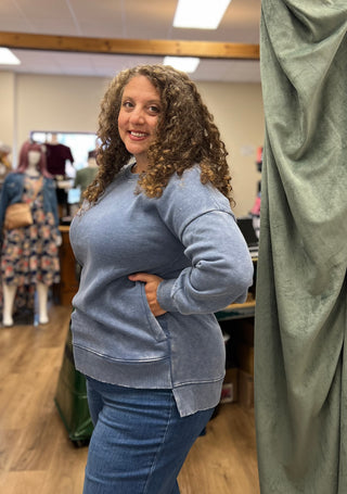 A woman with curly brown hair wearing a light blue crewneck sweatshirt and blue jeans, standing in a fitting room or boutique with a green curtain on the right.
