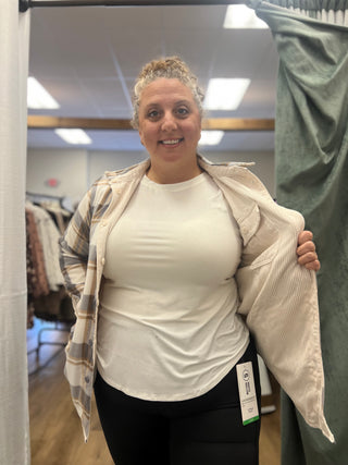 A woman stands in a fitting room wearing a white crewneck top, black leggings, and holding open a beige corduroy shirt over a plaid button-up shirt. A size tag is visible on the hem of the white top.