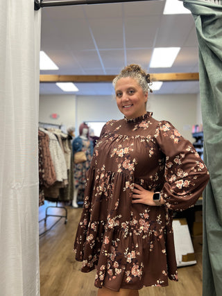 A woman wearing a brown long-sleeve tiered dress with a pink and beige floral pattern, standing in a fitting room.