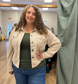 A woman with curly brown hair stands in an indoor fitting room or studio, wearing a light beige corduroy button-up shirt jacket over an olive green top and blue jeans.