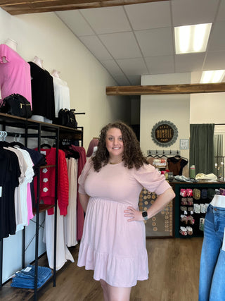 A woman with curly brown hair wearing a light pink long-sleeve ruffled dress, standing inside a clothing store next to racks of folded garments.