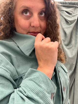 A close-up shot of a person wearing a sage green corduroy jacket, looking up and to the side with their hand near their chin, with a matching sage green fabric draped in the background.