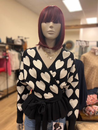A mannequin wearing a black wrap-style sweater with a white heart pattern, styled with a black belt and blue jeans in a clothing store.