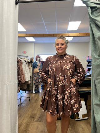 A woman modeling a brown floral tiered dress in a boutique fitting room, framed by white and green curtains on both sides.