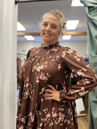 A woman wearing a brown long-sleeve floral dress with a ruffled high neck, standing in a fitting room or boutique.