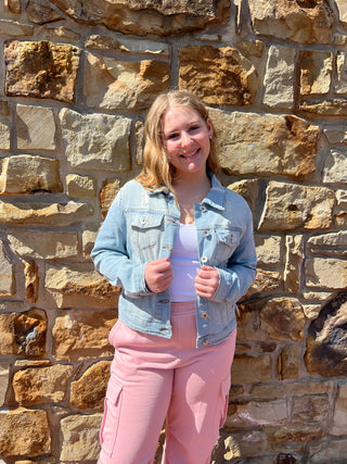 A young woman lying on a stone wall wearing a light blue denim jacket and pink pants, holding a clear plastic cup.