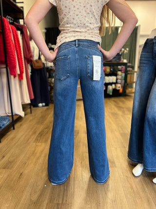 A person standing in a clothing store, viewed from the back, wearing blue wide-leg jeans with a visible white size tag on the back pocket. The store has wood floors, racks of colorful clothing, and other jeans hanging nearby.