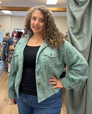 A woman with curly brown hair wearing a sage green corduroy utility jacket over a black top and blue jeans, standing inside a clothing store next to a green garment hanging on a stand.