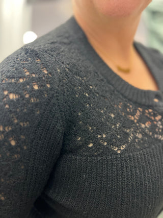 Close-up of a person wearing a dark charcoal gray knit sweater with an openwork lace pattern across the chest and shoulder.