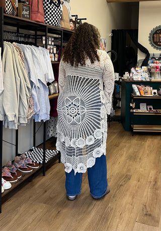 A woman viewed from behind wearing a white crochet cardigan with a large circular mandala pattern on the back, standing inside a retail store with clothing racks and wood flooring.