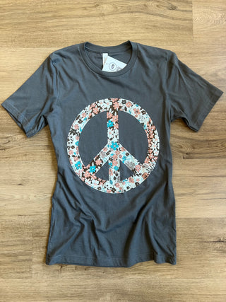 A dark gray short-sleeve t-shirt laid flat on a light brown wood floor, featuring a large peace sign graphic on the front filled with a floral pattern in pink, blue, and white.