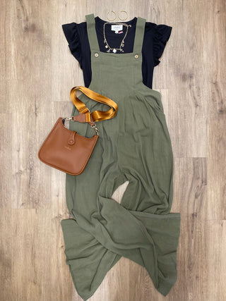 An olive green wide-leg overall jumpsuit styled in a flat lay with a black ruffled short-sleeve top, a brown leather crossbody bag, and gold jewelry, all arranged on a light brown wood floor.