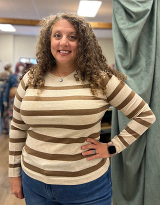 A woman with curly brown hair wearing a beige and brown horizontally striped long-sleeve knit top and blue jeans, standing in a fitting room with a green curtain in the background.