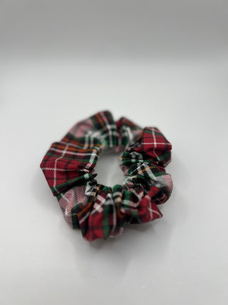 A fabric hair scrunchie featuring a red, green, and white tartan plaid pattern, photographed on a neutral light gray background.