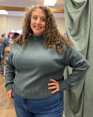 A woman with curly brown hair wearing a ribbed knit turtleneck sweater in a muted sage green, paired with blue jeans, standing in what appears to be a garment or fabric workshop.