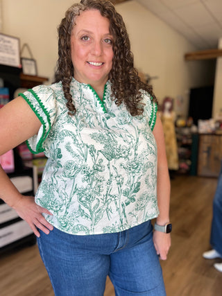A woman with curly brown hair wearing a green and white toile-print short-sleeve blouse with green trim, paired with blue jeans, standing in a shop interior.