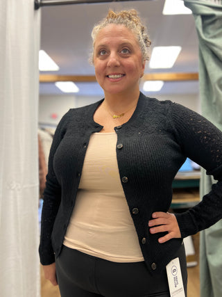 A woman wearing a black button-up cardigan over a beige tank top and black leggings, standing in a fitting room or boutique.
