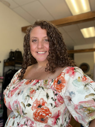 A smiling woman with curly brown hair wearing a white off-the-shoulder blouse with a vibrant pink, red, and orange floral print, photographed in a retail store setting.
